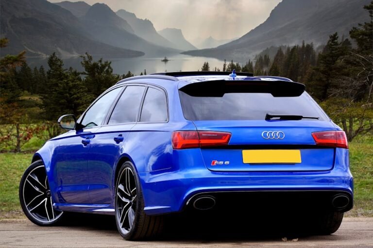 A sleek blue Audi RS 6 wagon parked against a stunning backdrop of Scottish hills and a serene lake.