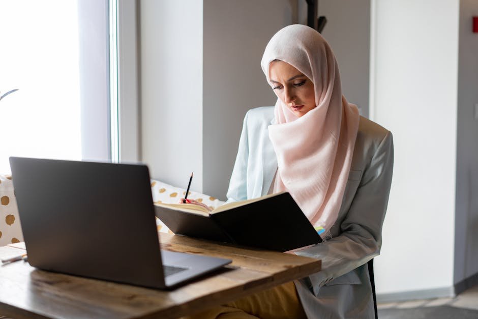 Focused woman in hijab reading a book at a wooden table with a laptop nearby.