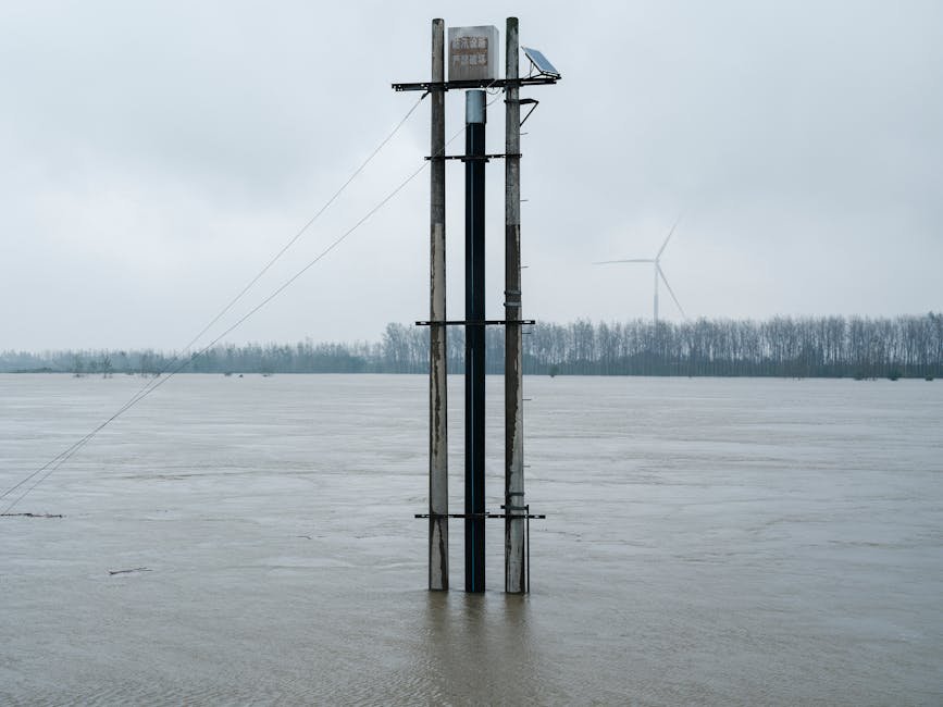 Flooded river landscape featuring a solar panel tower and distant wind turbine under overcast skies.