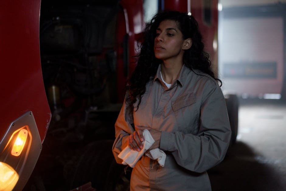A young female mechanic in workwear stands thoughtfully beside a vehicle in a dimly lit garage.