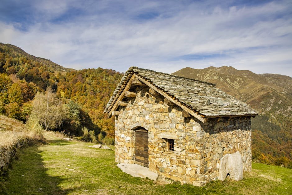 A rustic stone cabin nestled in a colorful autumn mountain landscape under a blue sky.