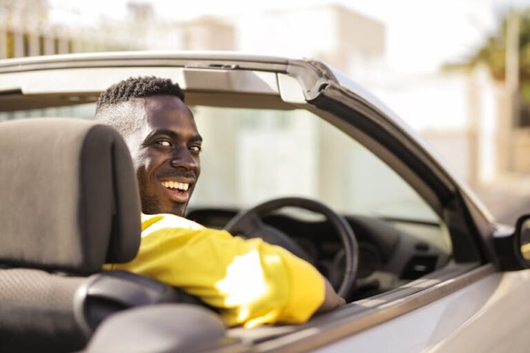 A man in a yellow shirt enjoys a sunny day while driving a convertible car.
