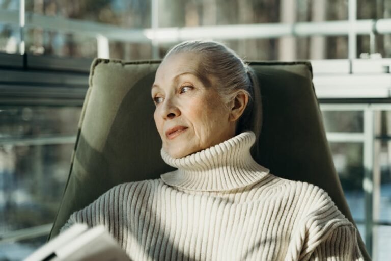 Elderly woman in a turtleneck sweater sitting comfortably indoors, looking pensive.