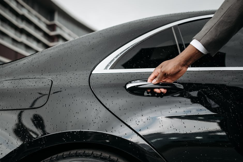 A close-up of a hand opening a black car door covered in raindrops, showcasing elegance and weather detail.