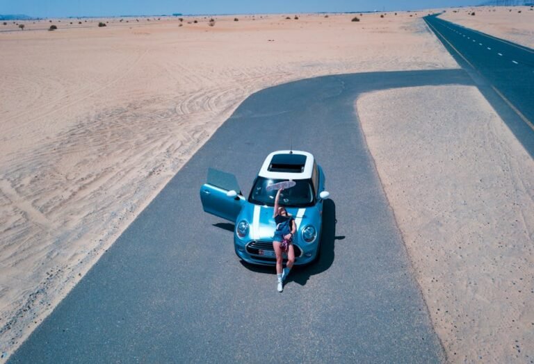 Woman with Mini Cooper on desert road near Dubai. Aerial shot captures vast sandy landscape.